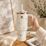 Hand holding a white Stanley tumbler with gold accents on a wooden surface.