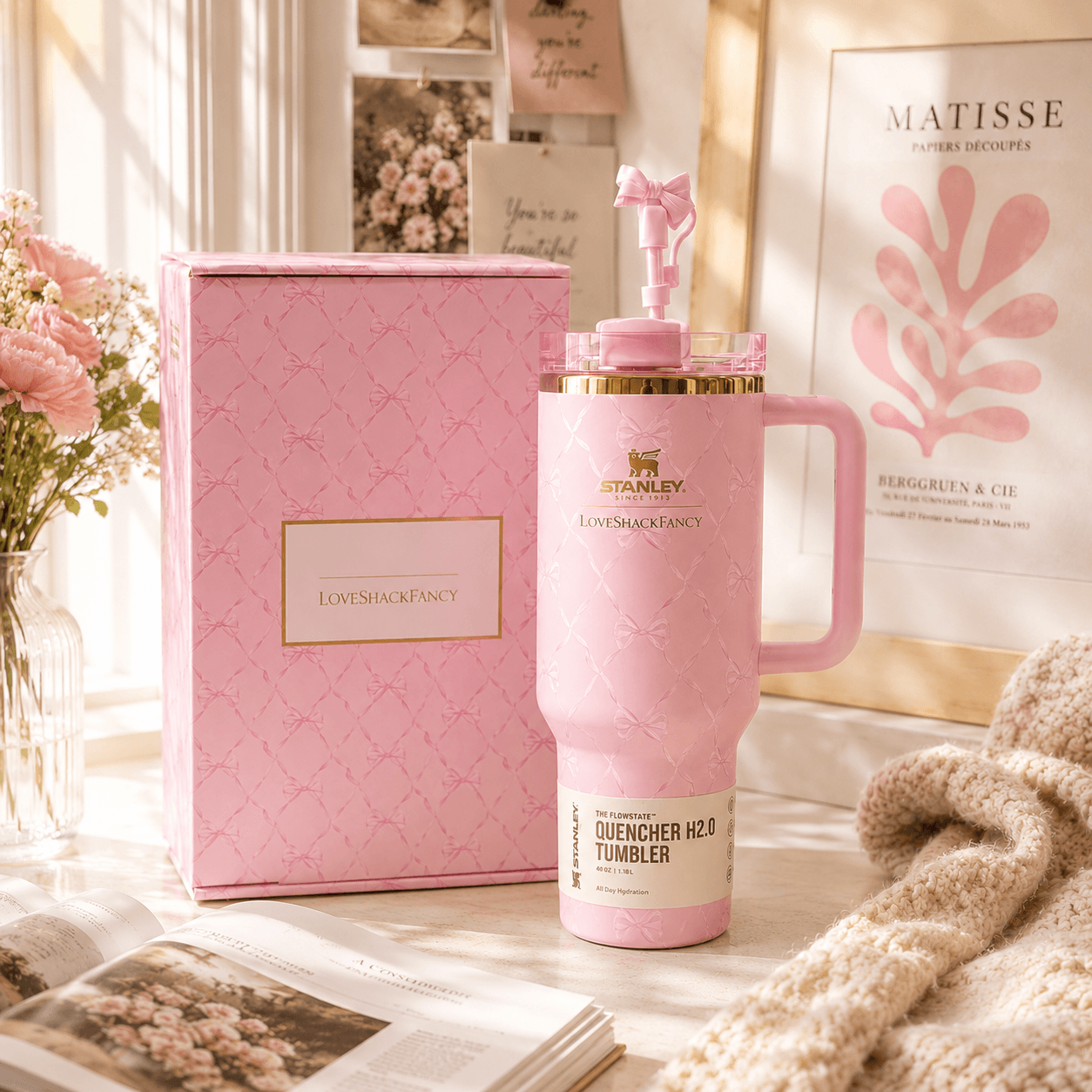 Pink tumbler with gold accents on a surface with a pink box and decorative items in the background.